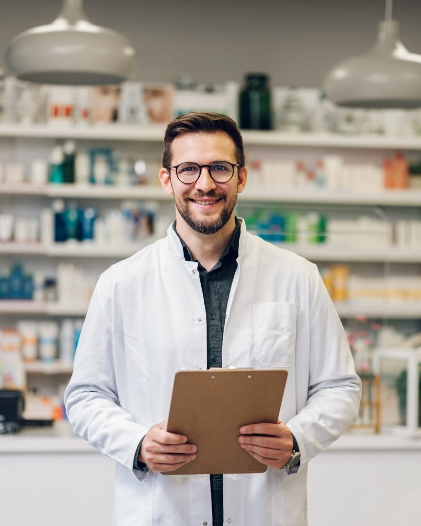 A young pharmacy intern doing inventory at the pharmacy shop as a part of their internship. He wears glasses and a white pharmacy coat. He is in front of a counter. Blurry background with shelves.